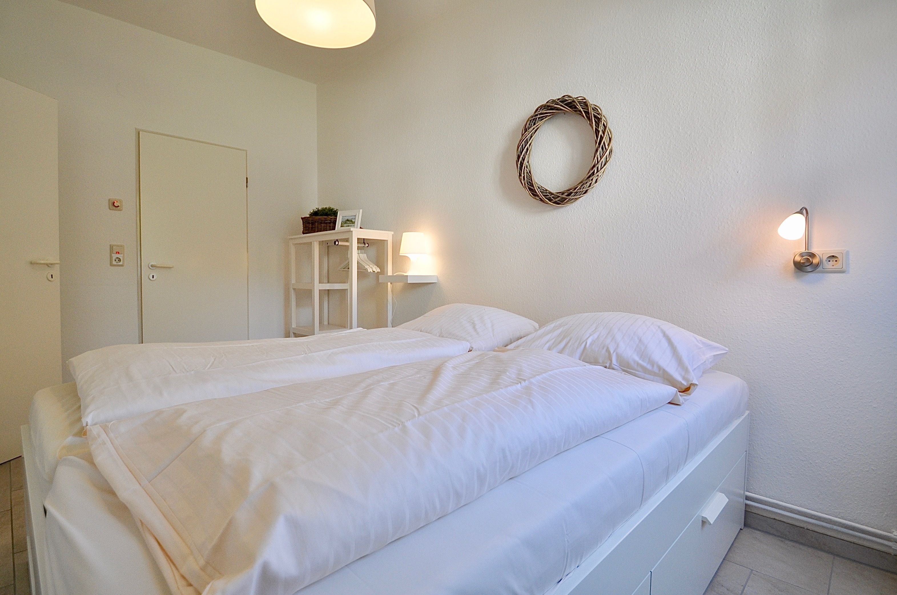 Uma cama de casal bem arrumada com roupa de cama branca, uma pequena prateleira com uma cesta e moldura de foto, uma luminária fixada na parede e uma coroa decorativa.
