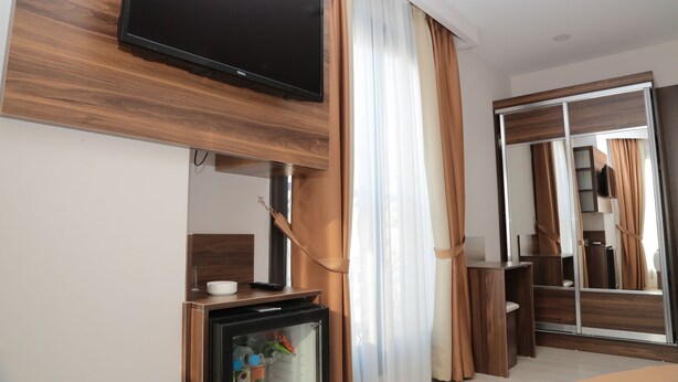 A modern hotel room with a flat-screen TV mounted on a wooden panel, a mini-fridge, a mirror, and a wardrobe with sliding doors. - Paris Prestige (Izmir)