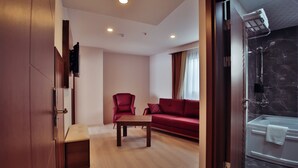 A hotel room with a wooden floor, a red sofa, a red armchair, a wooden coffee table, a TV mounted on the wall, and a shower area with a marble wall. - Paris Prestige (Izmir)