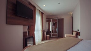 A hotel room with a large bed, a flat-screen TV mounted on the wall, a wooden wardrobe, a mirror, and a seating area with a chair and a small table. - Paris Prestige (Izmir)