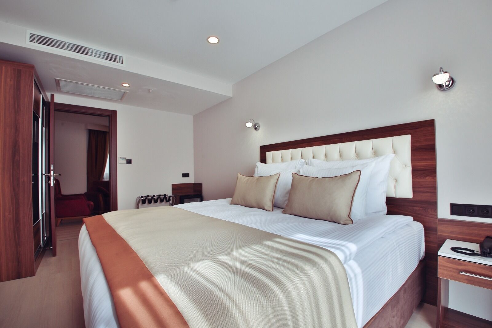 A hotel room with a large bed, a wooden headboard, a bedside table, a wardrobe, and a door leading to another room.