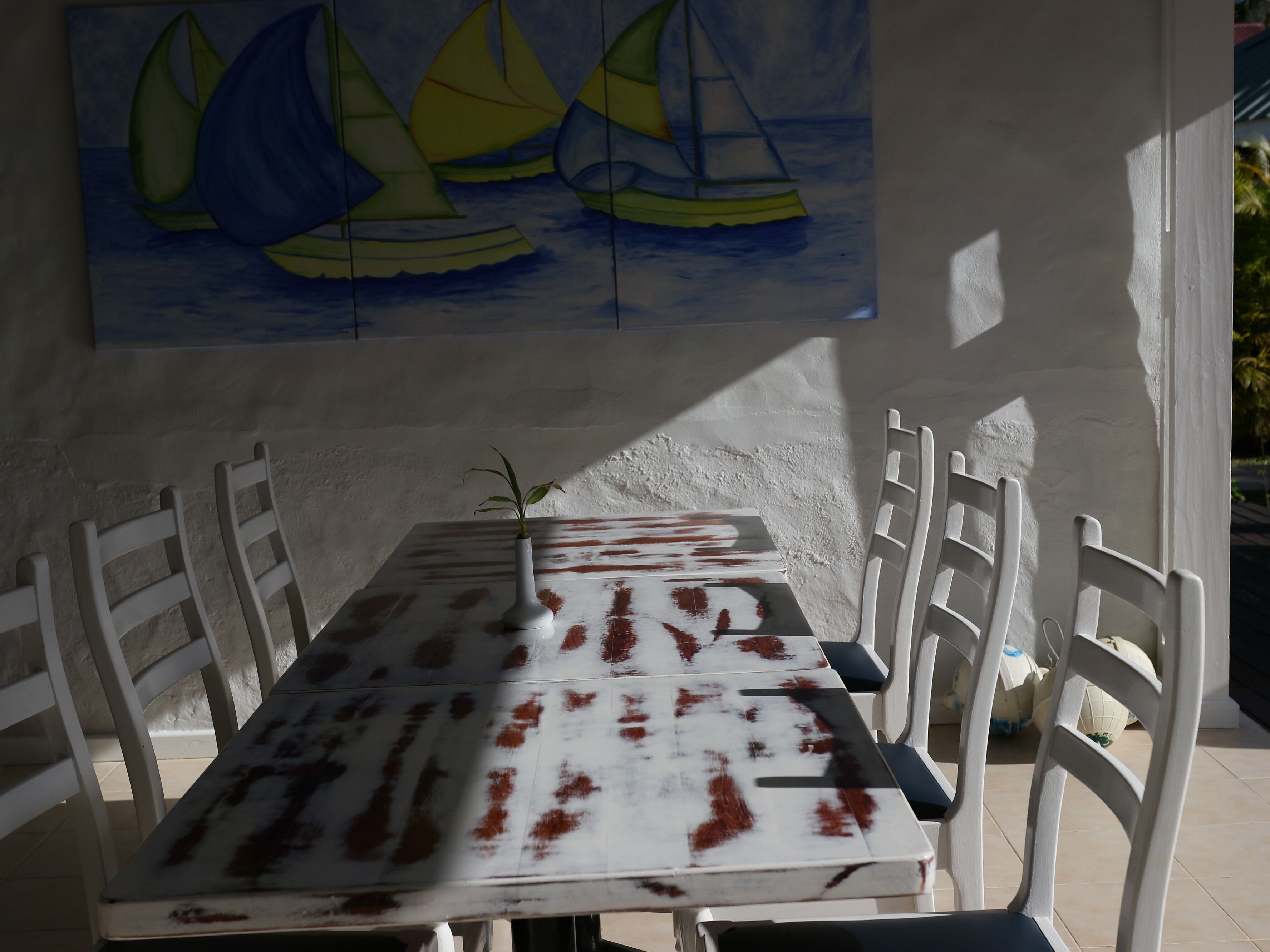 Una mesa de comedor con un diseño marmolado blanco y rojo, rodeada de sillas blancas, frente a una pared blanca con una pintura de un velero.