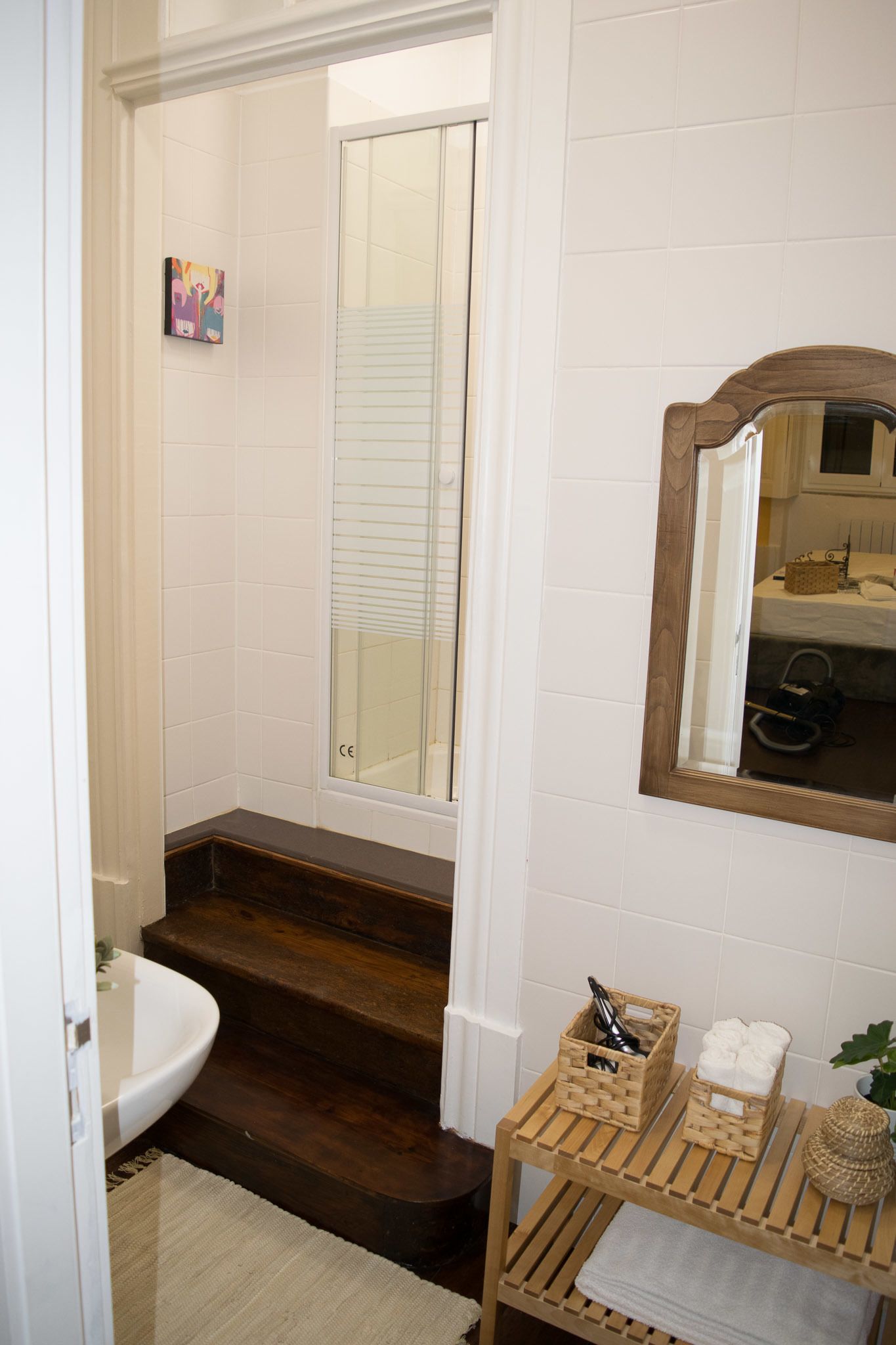 Un baño con paredes de azulejos blancos, suelo de madera, un lavabo blanco, un espejo y un estante de madera con toallas y cestas.