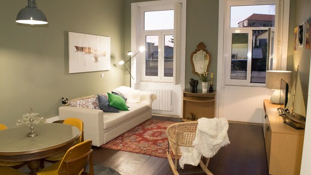 A cozy living room with a white sofa, a dining table, and a rocking chair. There are two windows providing natural light, and a mirror hangs on the wall. - Merc Porto Central (Porto)
