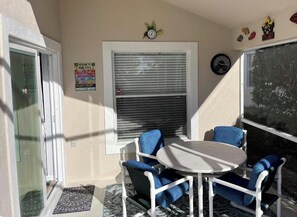A small outdoor patio with a round table and blue chairs, a closed window with blinds, and a door with a glass panel.