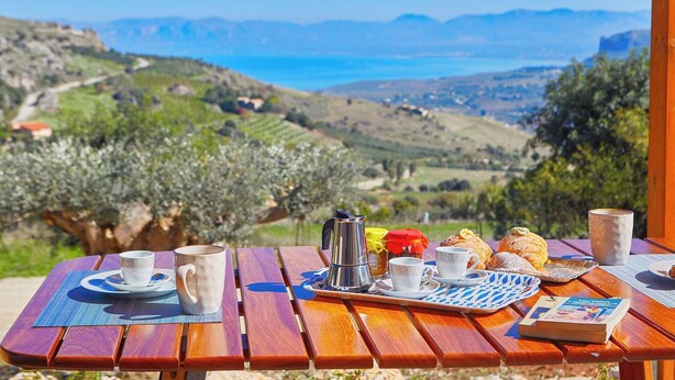 A wooden table set with breakfast items and a book, overlooking a scenic landscape with hills and a body of water.