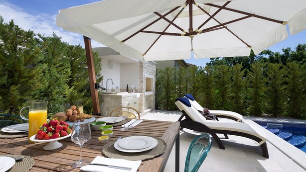 Un área de comedor al aire libre con una mesa de madera preparada para dos personas, una sombrilla blanca y una silla de descanso junto a la piscina.