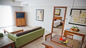Ein Hotelzimmer mit einem grünen Sofa, einem Holztisch mit Obst-Dekoration, einem Fernseher und einem Bett mit einem roten Kissen.