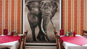 Un comedor con dos mesas, cada una cubierta con mantel blanco y camino de mesa rojo. Hay sillas alrededor de las mesas, y un cuadro enmarcado con un elefante destaca en la pared.