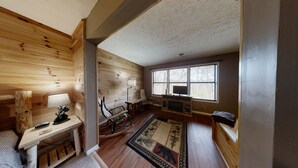 A cozy cabin interior with wooden walls, a fireplace, a desk with a lamp, a rocking chair, and a large window with blinds.