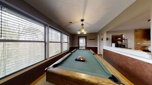 A pool table in a room with large windows, a ceiling fan, and a reception area with a wooden finish.