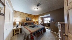 A cozy bedroom with a wooden headboard, a large bed with a bear-themed quilt, a nightstand with a lamp, and a window with blinds.