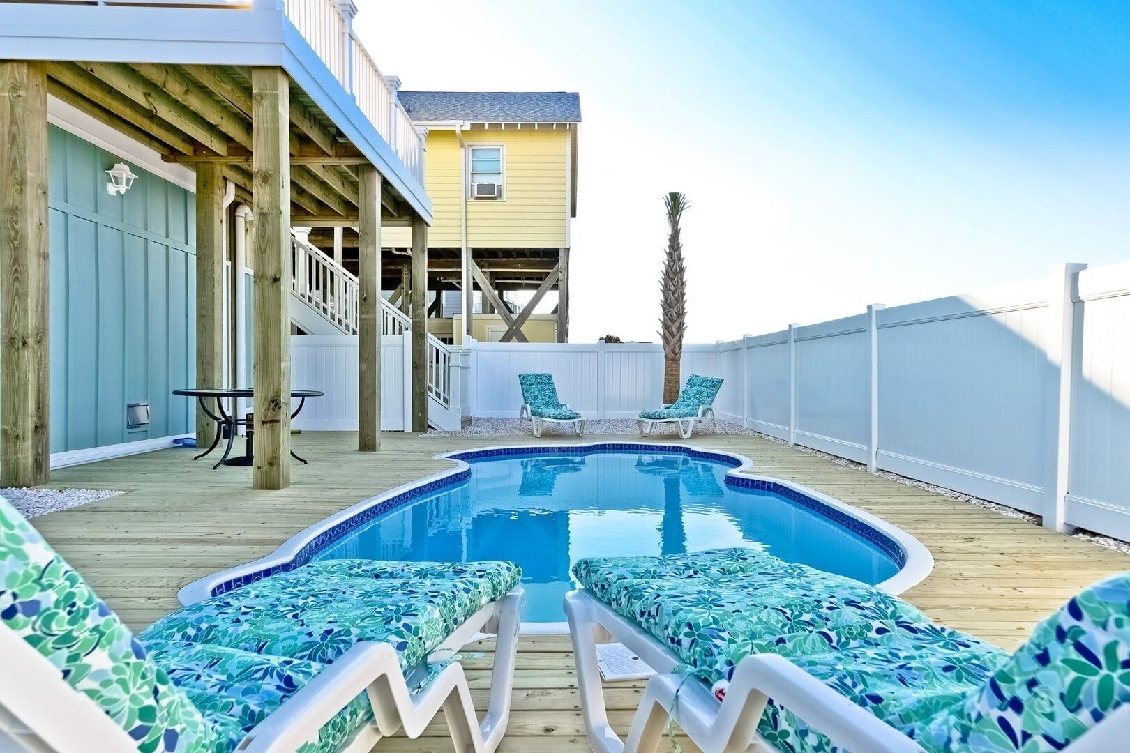 A swimming pool with lounge chairs, a wooden deck, a white fence, and a building with a balcony.