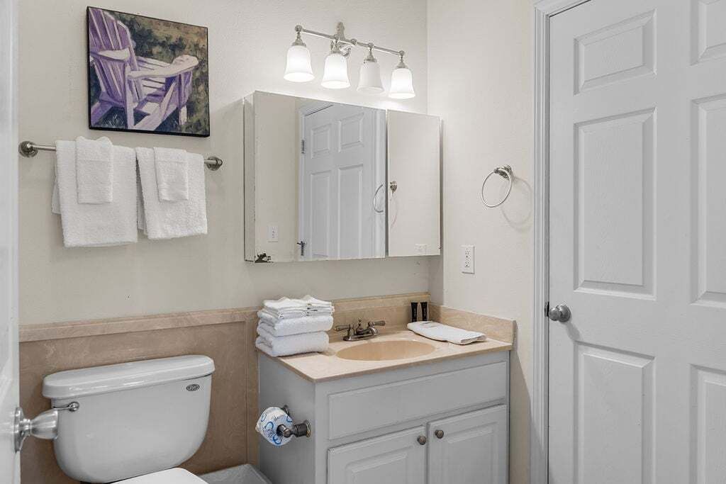 A bathroom with a toilet, sink, and mirror. There are towels on the wall and a painting above the mirror.