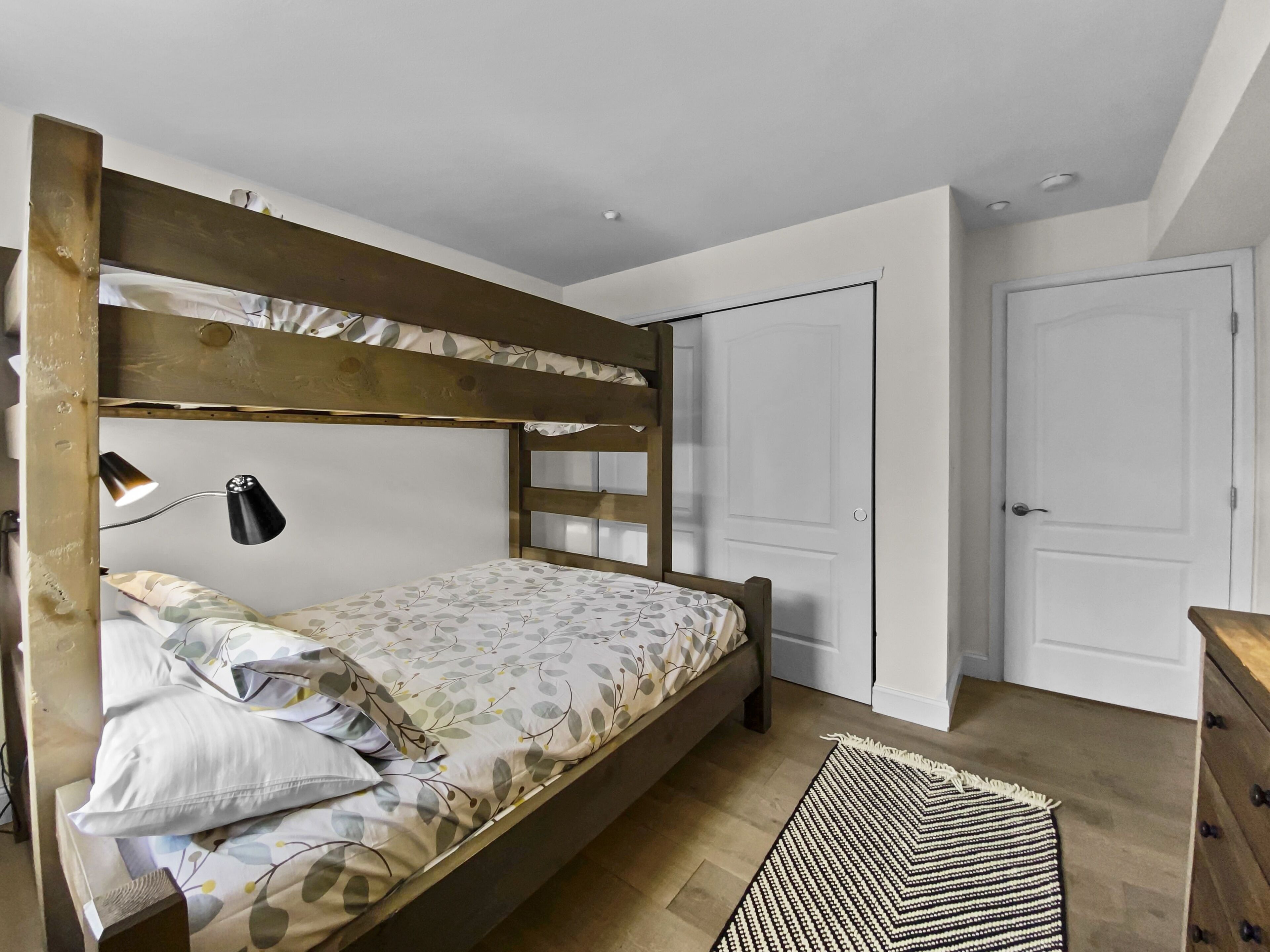 Een stapelbed met een houten hoofdbord, een bedlampje en een geborduurde spreideken. Er staat een houten dressoir en een deur in de kamer.