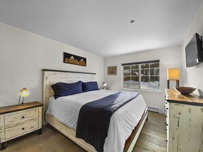 A bedroom with a bed, a nightstand, a dresser, a window with a view of a snowy landscape, and a mounted TV.