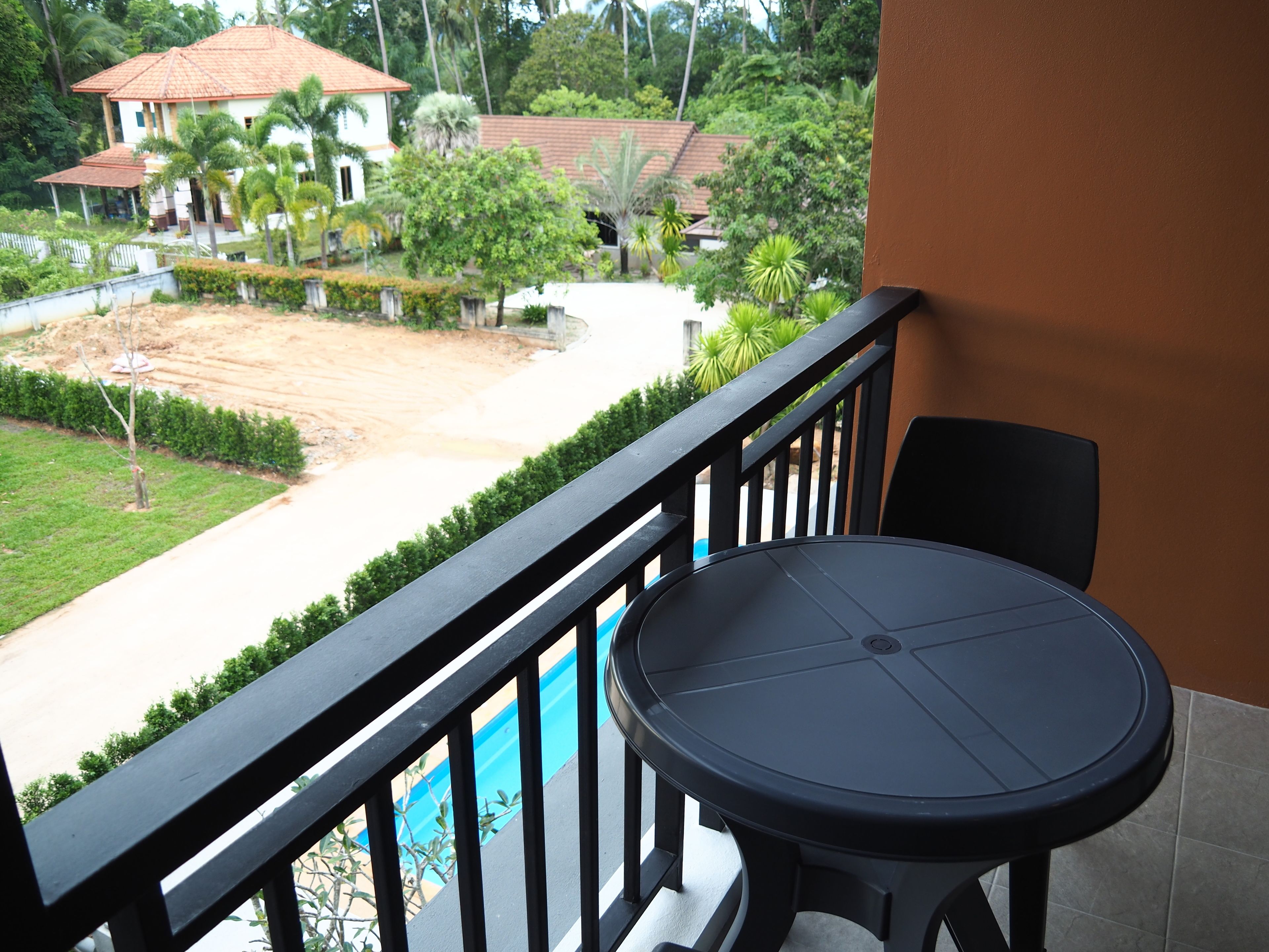 A balcony with a round table and chairs, overlooking a landscaped area with a dirt path, greenery, and a building with a red roof.