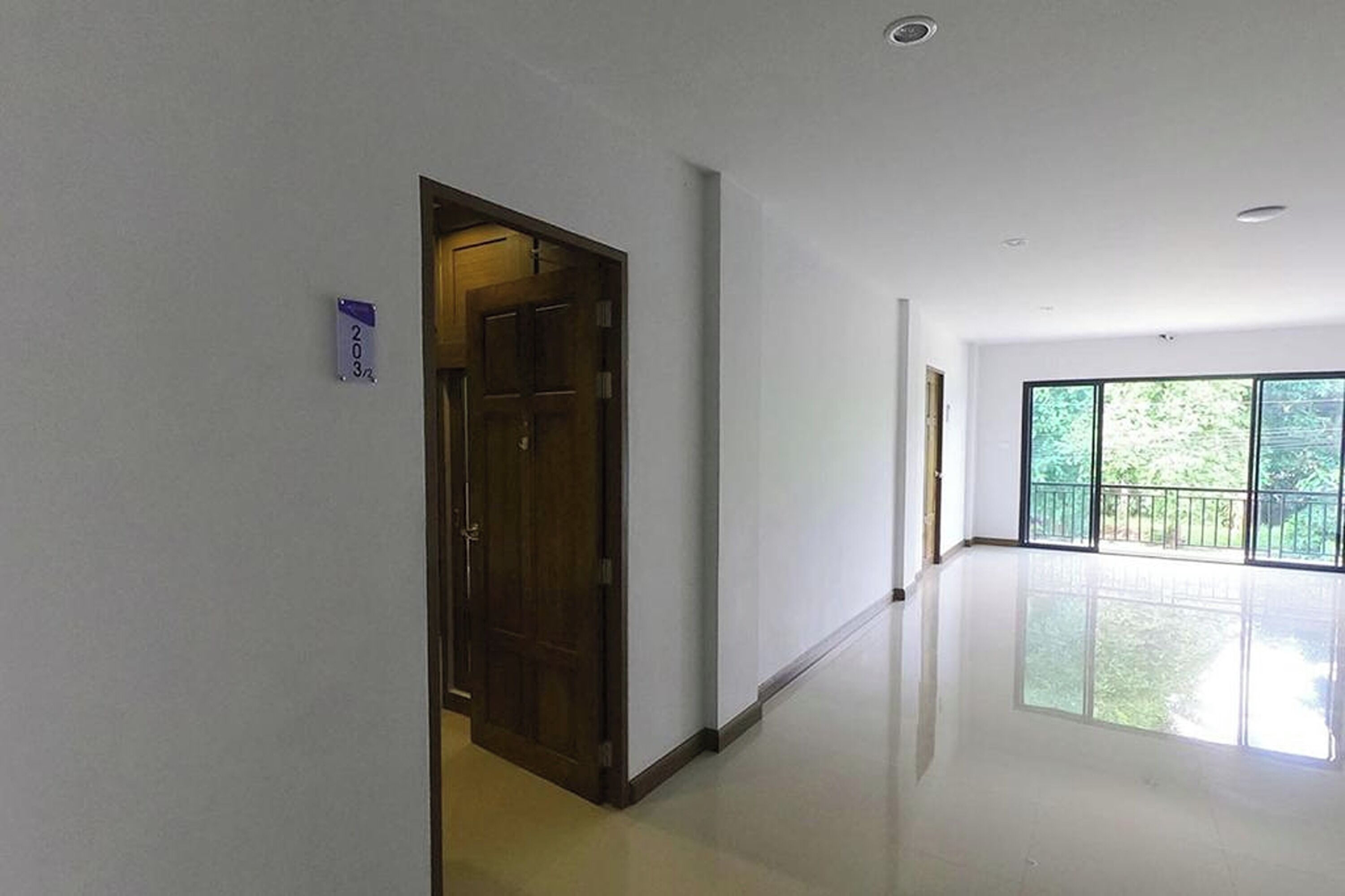 A hallway with a wooden door, a number sign, and large windows with a view of greenery.