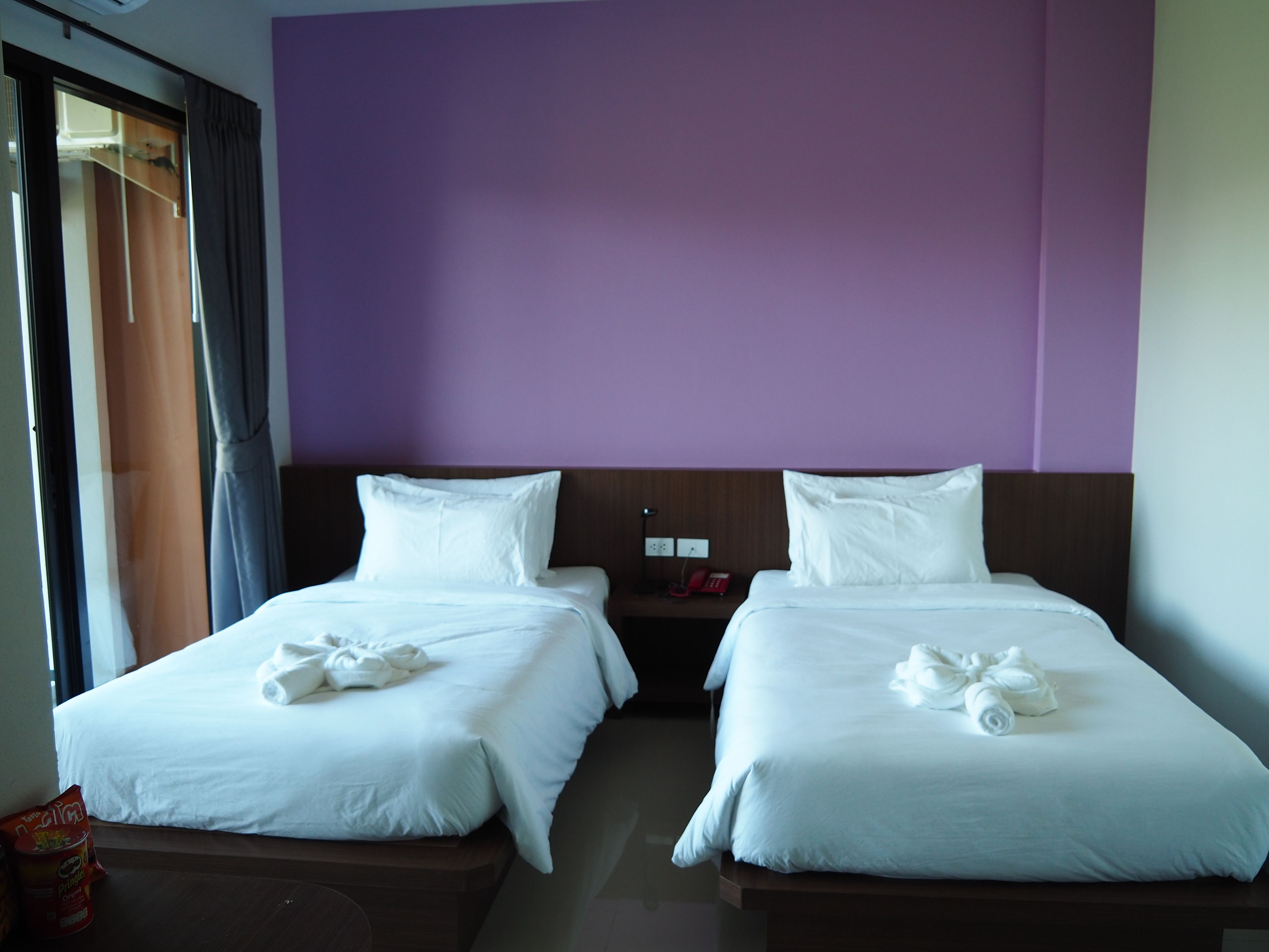 Una habitación de hotel con dos camas, cada una con ropa blanca y toallas, una pared de acento morada y una mesita de noche pequeña con un teléfono rojo.