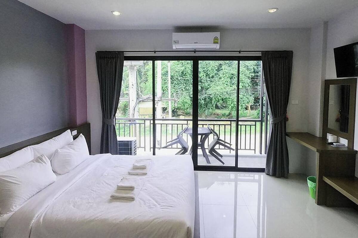 A hotel room with a bed, a TV, a balcony with a table and chairs, and a view of greenery outside.