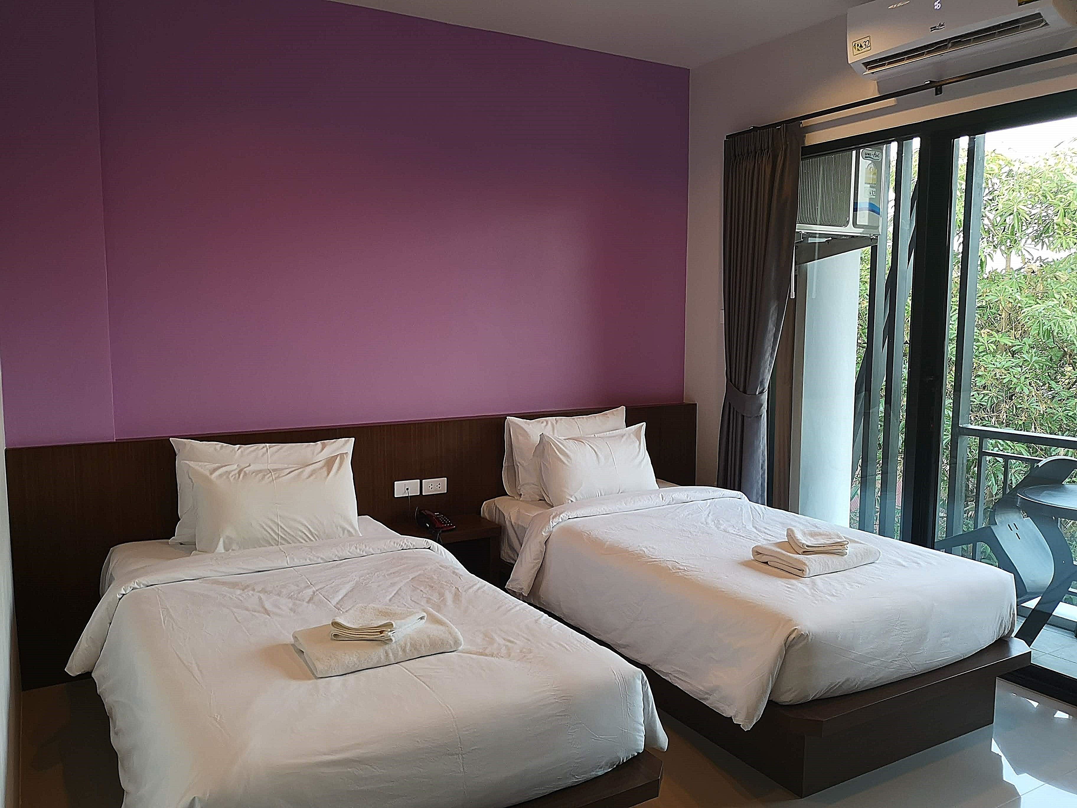 A hotel room with two beds, purple walls, and a balcony with a view of greenery.