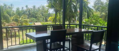 A balcony with a table and chairs, overlooking a lush garden with palm trees and a clear sky.