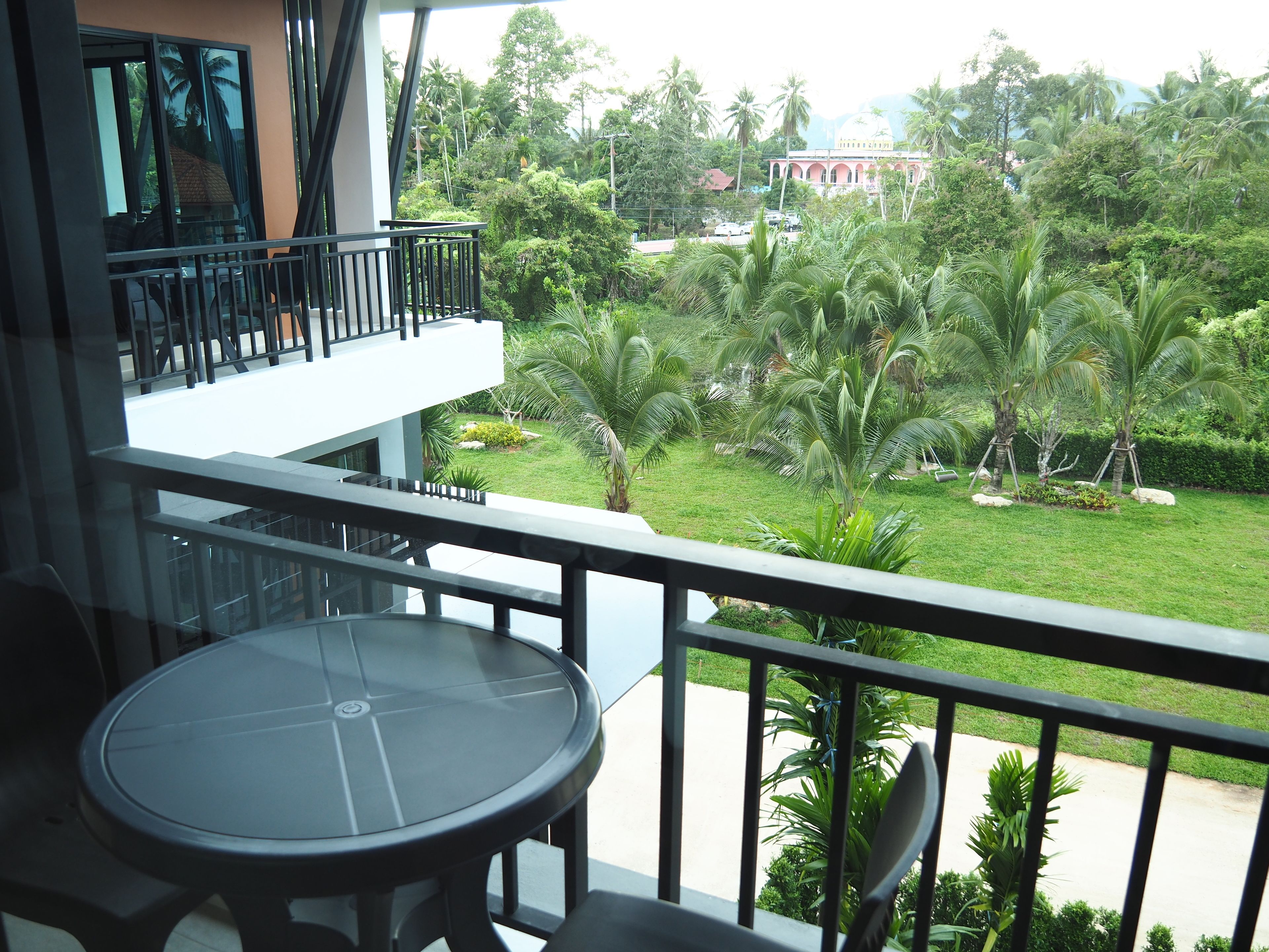 A balcony with a table and chairs, overlooking a lush garden with palm trees and a building in the background.