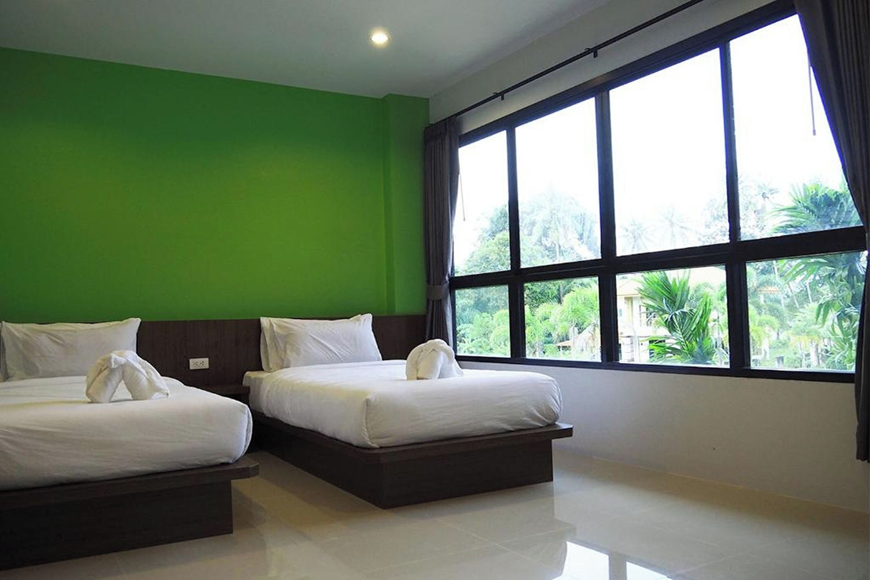 A hotel room with two beds, a green wall, and large windows with a view of greenery outside.