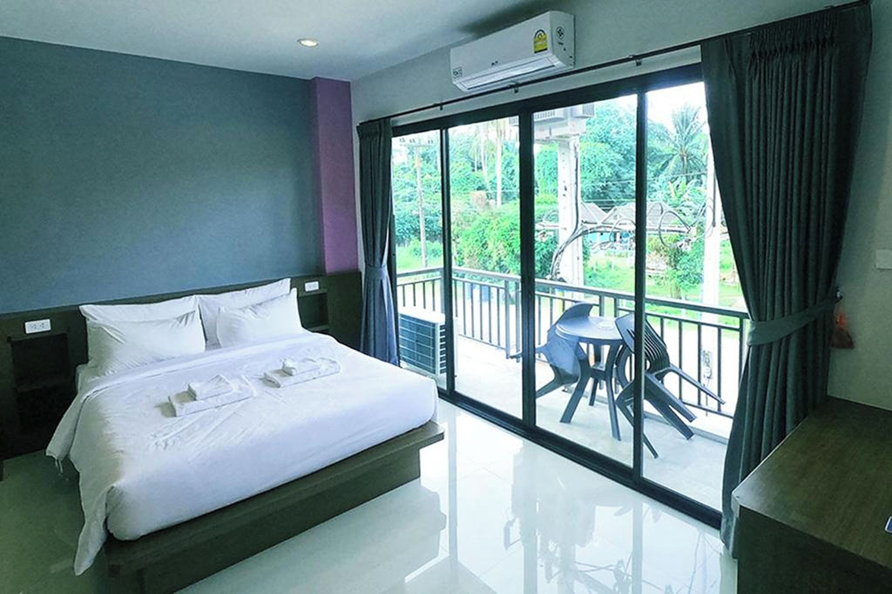 A hotel room with a bed, a balcony with chairs, and a view of greenery.