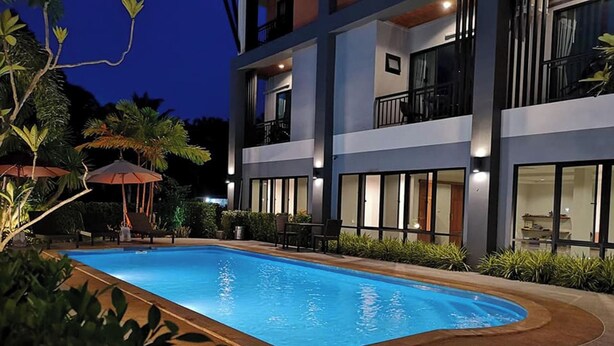 Uma piscina com borda curva, rodeada por uma área ajardinada com plantas e um espaço de estar com guarda-sóis.