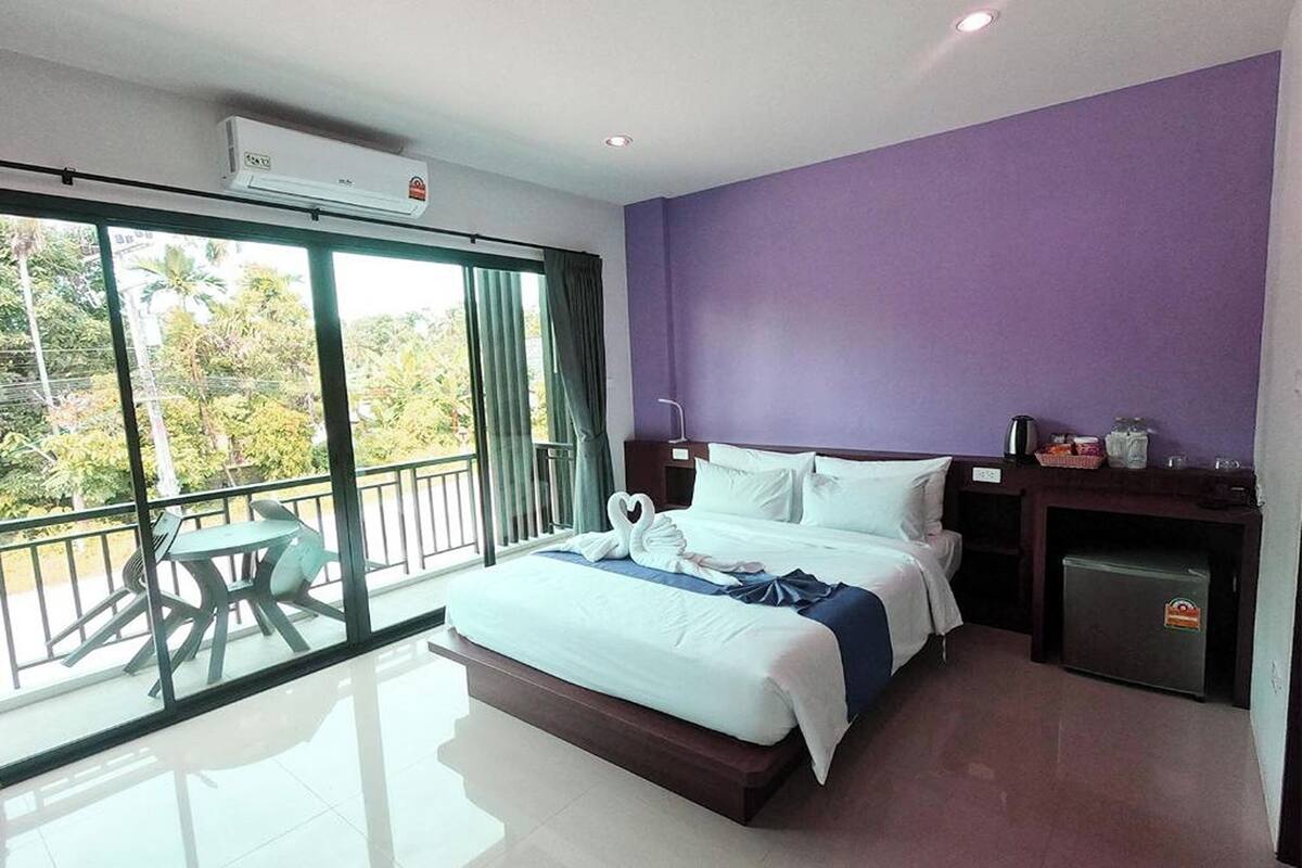 A hotel room with a large bed, a small desk, a refrigerator, and a balcony with a table and chairs.