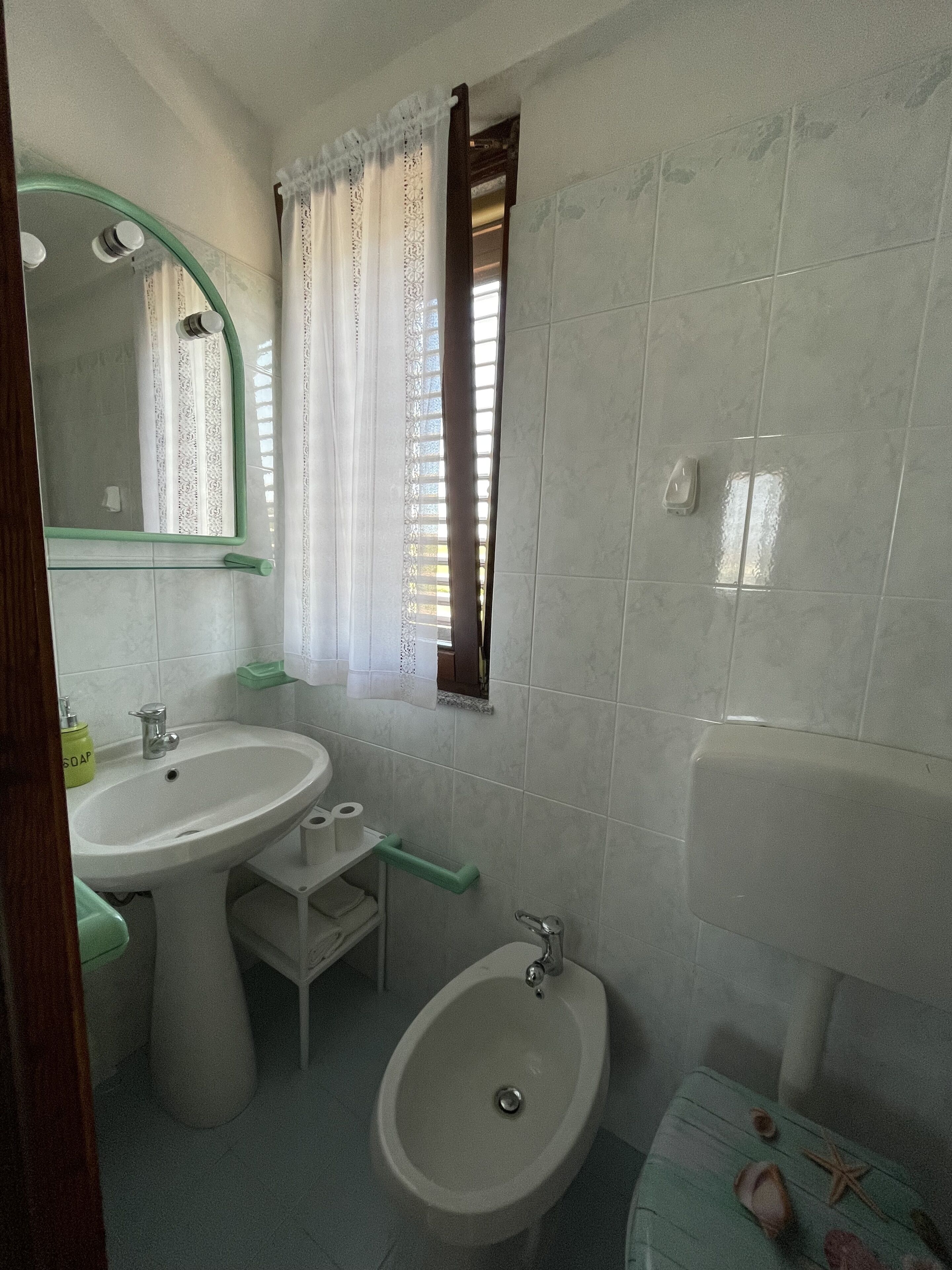 A bathroom with a sink, bidet, and bathtub. There is a window with curtains, a mirror, and a soap dispenser.