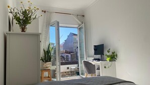 A bedroom with a bed, a desk with a computer, a chair, and a view of buildings outside.