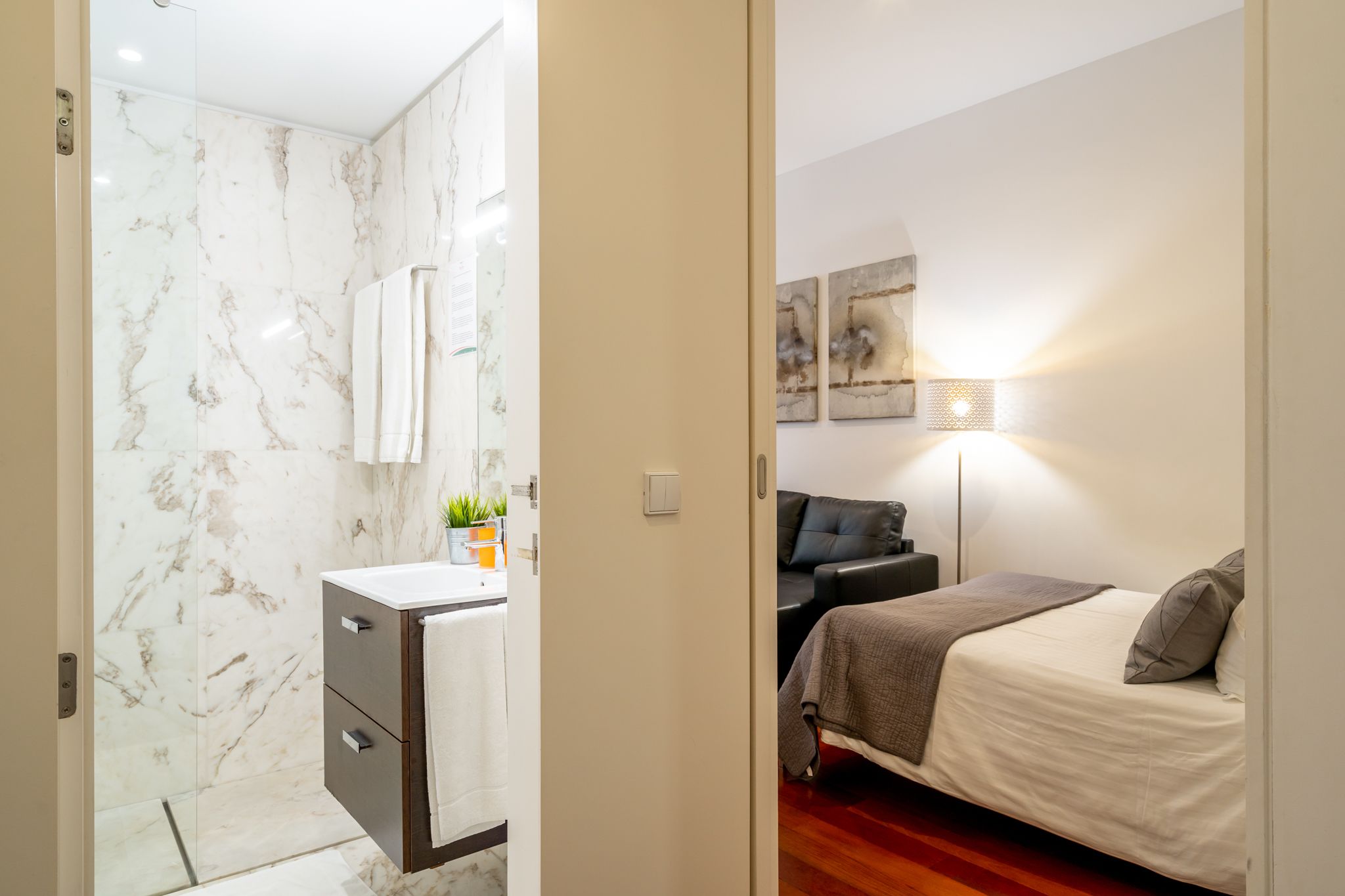Un baño moderno con pared de mármol y un dormitorio independiente con cama, mesita de noche y lámpara.
