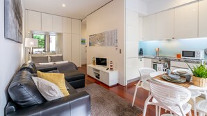 Uma sala de estar moderna com um sofá de couro preto, um espaço de jantar com cadeiras brancas e uma cozinha com armários brancos e um micro-ondas.