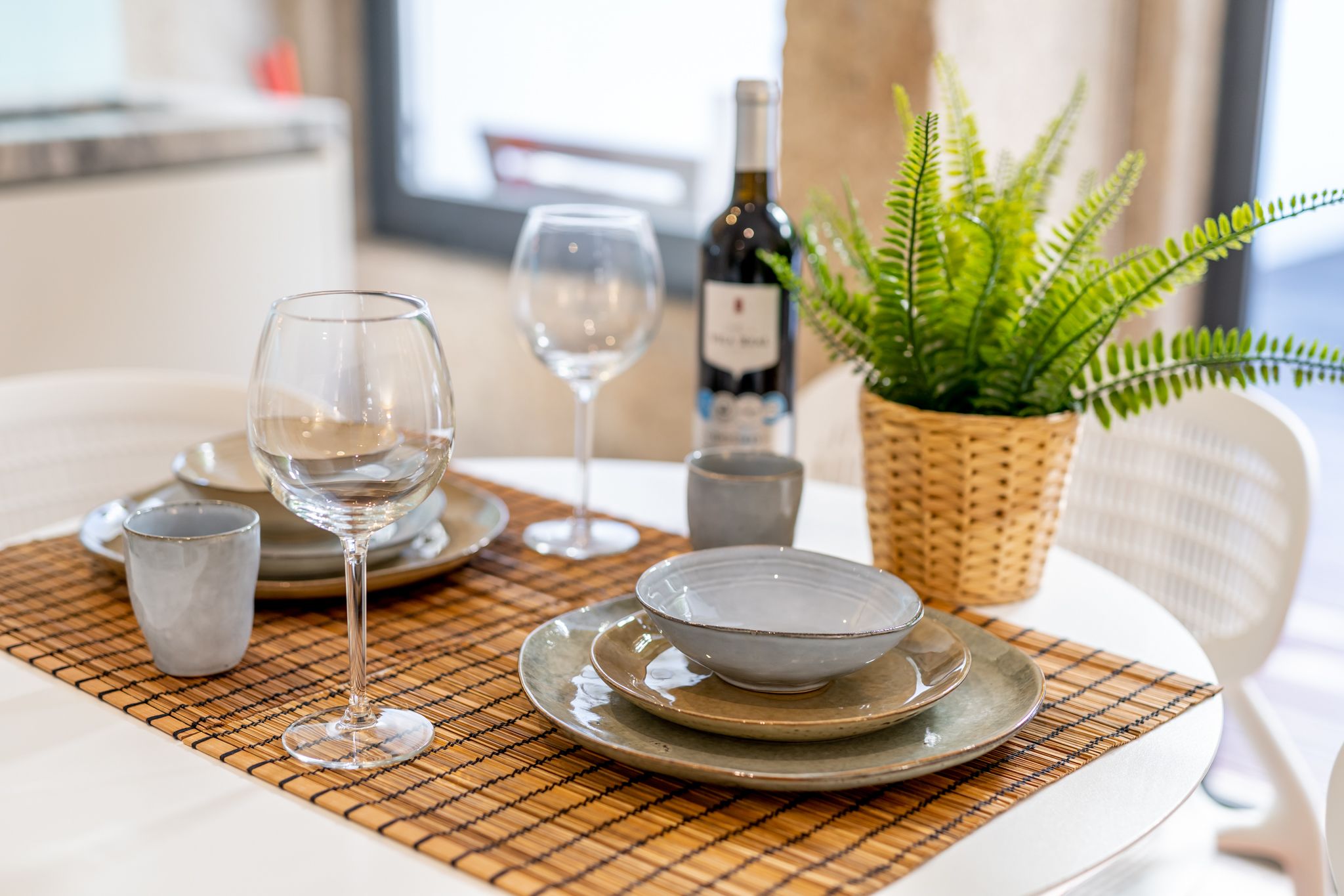 Een eettafel met borden, kommen en wijnglazen op een geweven placemat. Ook een fles wijn en een potplantje met een varen zijn zichtbaar.