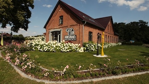 A brick building with a sign reading "KINSTRAILS BASCHHORST" surrounded by well-maintained gardens and a clear sky.