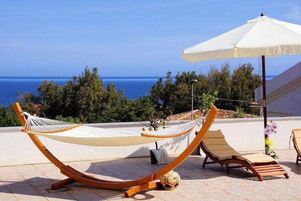 A hammock and a lounge chair on a tiled patio with a view of the sea.