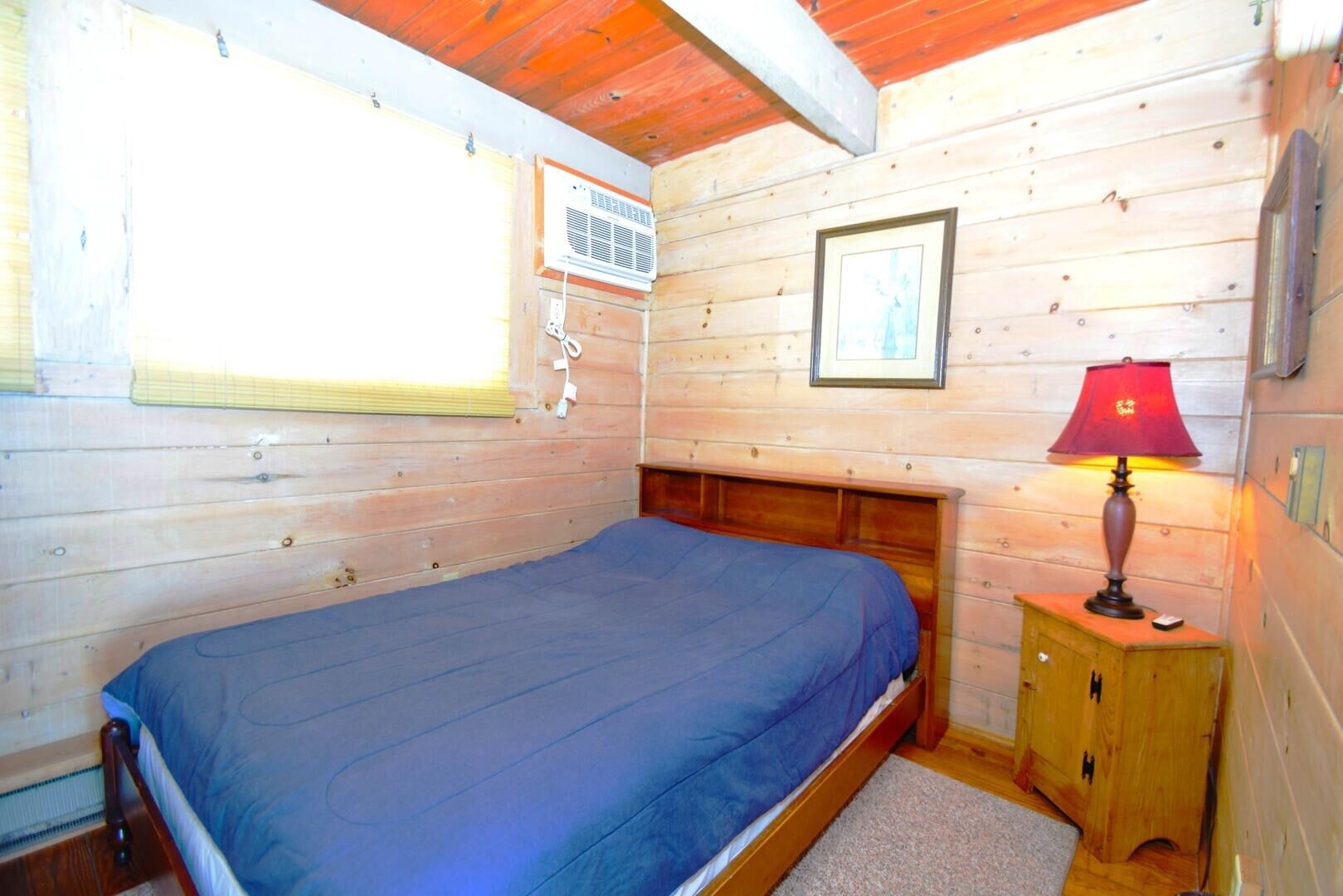 A wooden cabin room with a bed, a window with blinds, an air conditioning unit, a framed picture, and a bedside lamp.