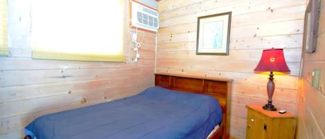 A wooden cabin room with a bed, a window with blinds, an air conditioning unit, a framed picture, and a bedside lamp.