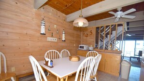 Een houten hut binnen met een eettafel, witte stoelen, een plafondventilator en een keuken met een magnetron.