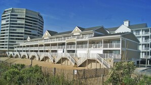 Een strandhotel met meerdere balkons, een zandstrand ervoor en een hoog gebouw op de achtergrond.