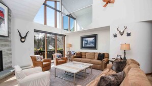 A spacious living room with a high ceiling, large windows, and a fireplace. There are multiple seating areas including a sofa, chairs, and a covered chair. - Ge Inn Plaza #109 4 Bedroom Condo by RedAwning (Vail)