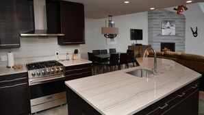 A modern kitchen with dark cabinetry, a large island with a sink, a gas stove, and a built-in oven. There is a dining area with a table and chairs, and a fireplace in the background. - Ge Inn Plaza #109 4 Bedroom Condo by RedAwning (Vail)