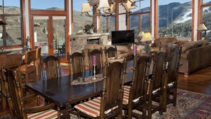 Un coin repas avec une grande table en bois, des chaises et une cheminée. On aperçoit un téléviseur et un canapé au fond.