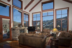 Ein geräumiges Wohnzimmer mit großen Fenstern, einem Kamin aus Stein, Ledersofas und einem Fernseher.