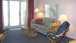 Ein Wohnzimmer mit einer Couch, zwei Sesseln, einem Glastisch und einer Stehlampe. Eine Glastür verschiebt sich zu einem Außenbereich.