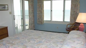 Ein Schlafzimmer mit Bett, Stuhl, Lampe und Meerblick durch ein Fenster.