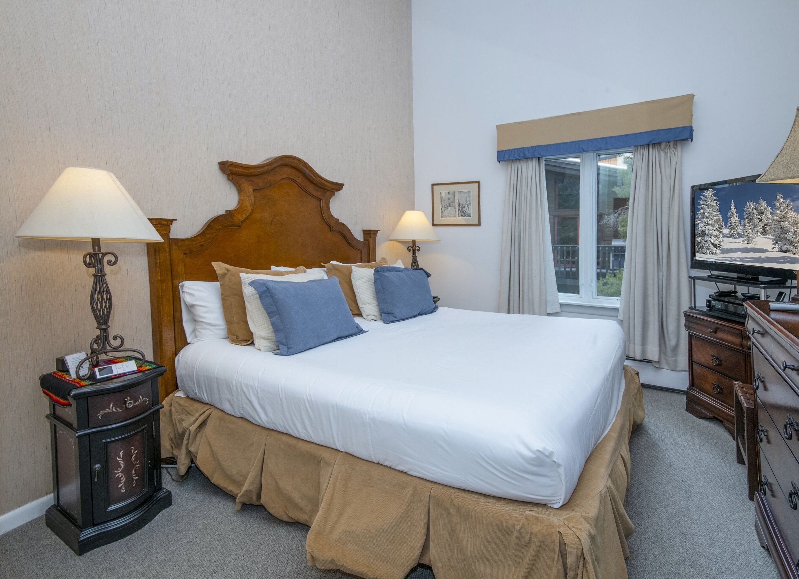A hotel room with a large bed, two bedside lamps, a television, a dresser, and a window with curtains.