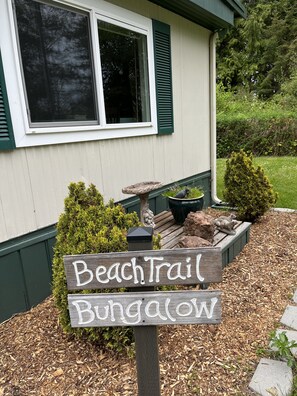 A wooden sign with the text "Beach Trail Bungalow" in front of a house with green shutters and a window.